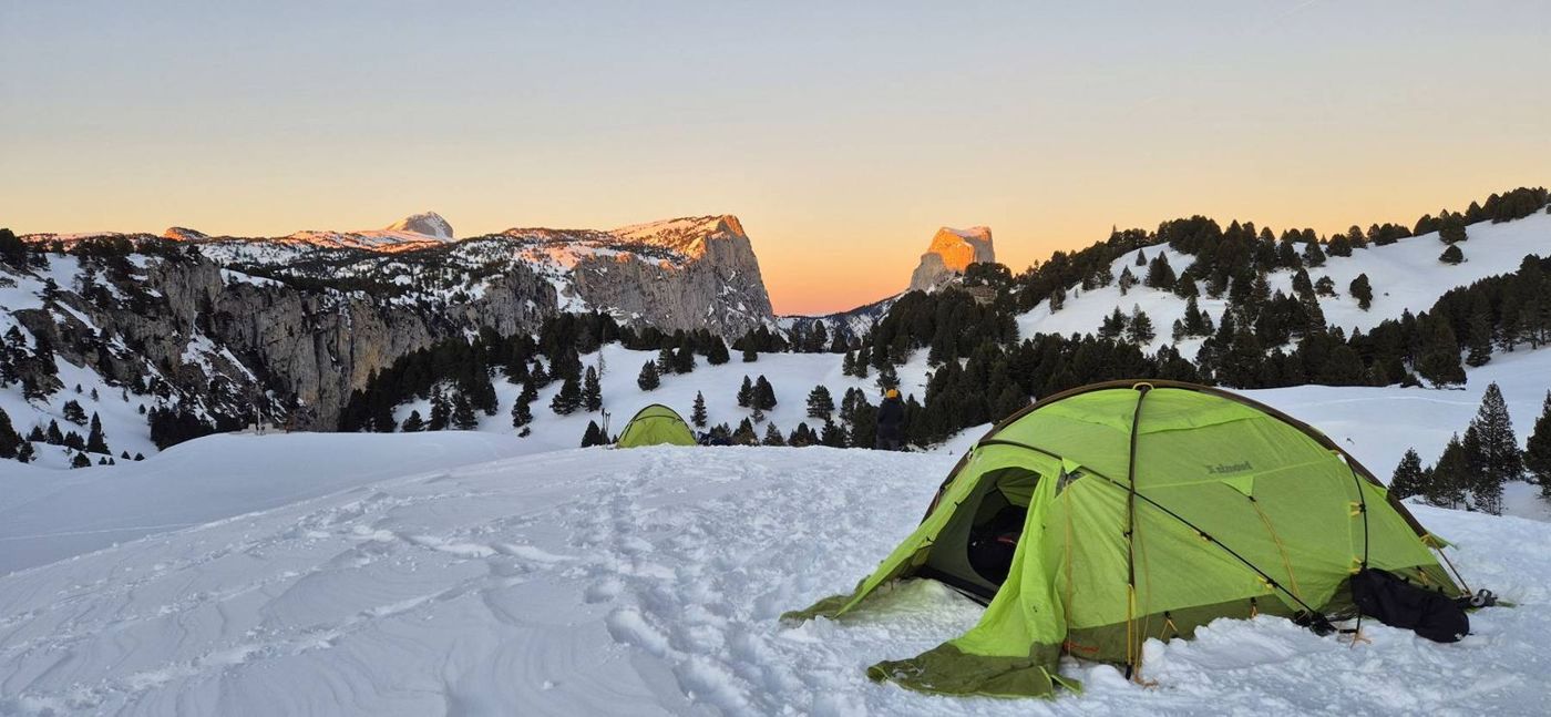 Une tente verte dans la neige au lever du jour, face aux falaises du Vercors
